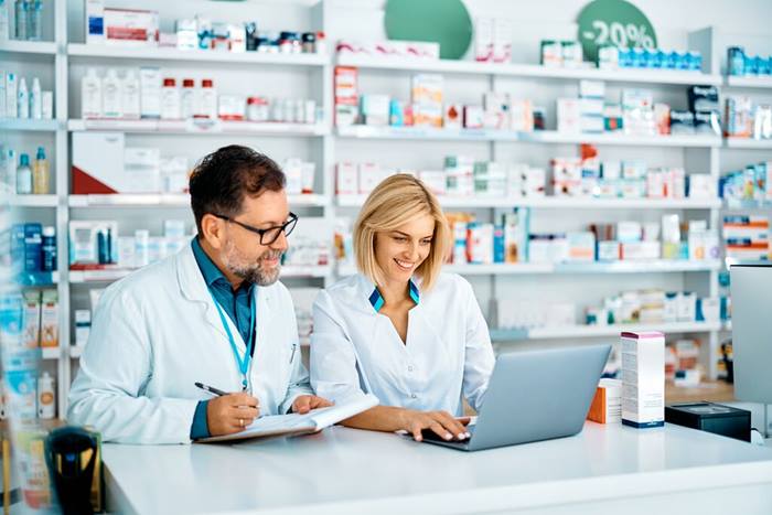 Pharmacist using laptop while colleague takes notes in pharmacy